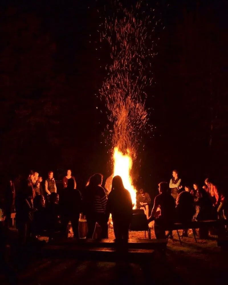 Una gran fogata de noche lanzando una columna de chispas al cielo, rodeada por una rueda de gente en silueta que conversa de pie y sentada en bancos de madera.