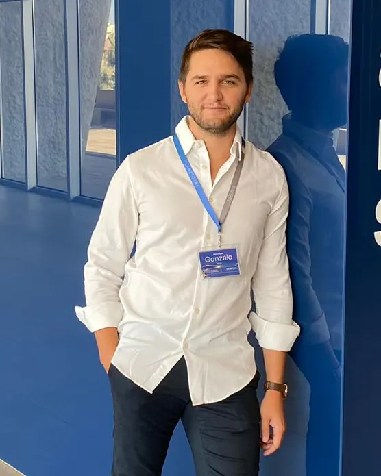 Gonzalo Rioja, de pie y con camisa blanca, sonríe a la cámara apoyado en un panel azul, con la acreditación de un evento colgada al cuello.